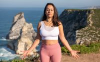 A woman in activewear practices meditation by the sea on a sunny day in Portugal.