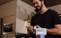Man in kitchen holding and reviewing a bottle of supplements, indicating health-conscious lifestyle.